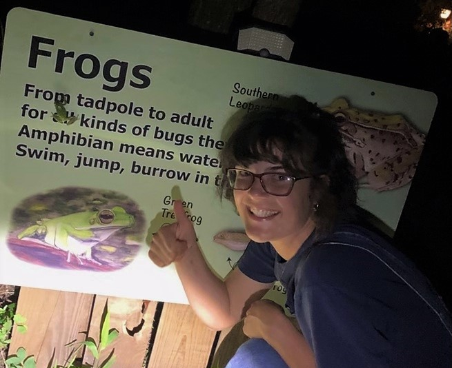 Abby Cervenka posing in front of a sign about frogs with a real frog on it!